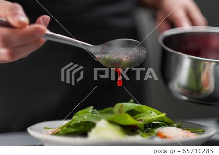 A female chef in a white uniform and a black apron in the restaurant kitchen. The cook pours red cranberry salad sauce. Making and decorating food. 65710285