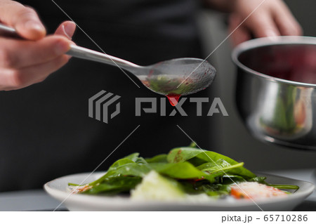 A female chef in a white uniform and a black apron in the restaurant kitchen. The cook pours red cranberry salad sauce. Making and decorating food. 65710286