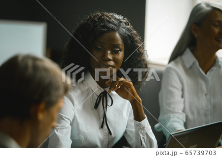 Diverse team brainstorming in office. Highlighted focus on African-American young woman sitting at table along with grey-haired mature woman and Caucasian man Diverse team brainstorming in office. Highlighted focus on African-American young woman sitting at table along with grey-haired mature woman and Caucasian man 65739703