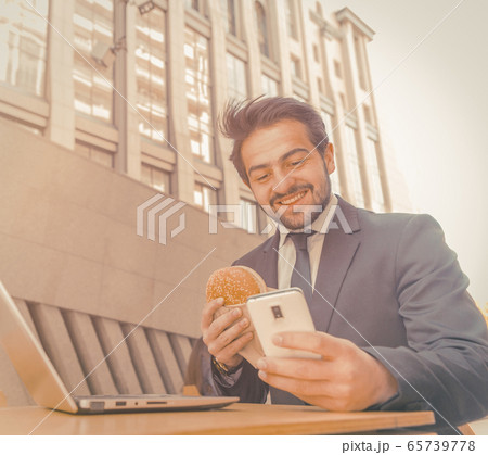Businessman eats in fast food restaurant typing on smart phone. Young man has a break sitting at table outdoors against background of beautiful city building. Business on the go concept. Toned image 65739778