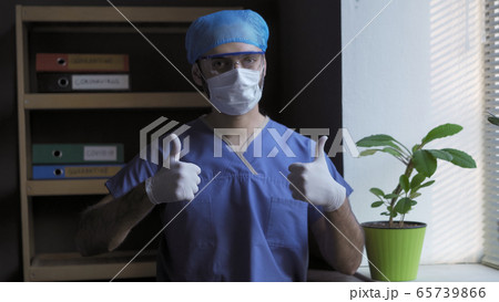 Doctor shows Thumbs up gesture. Positive gesture from male medic standing near the window against the background of a medical office. Pandemic ending concept 65739866