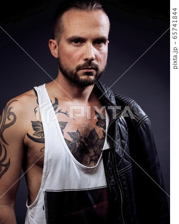 A man with tattooes on his arms. Silhouette of muscular body. caucasian brutal hipster guy with modern haircut, looking like criminal close up A man with tattooes on his arms. Silhouette of muscular body. caucasian brutal hipster guy with modern haircut, looking like criminal close up 65741844