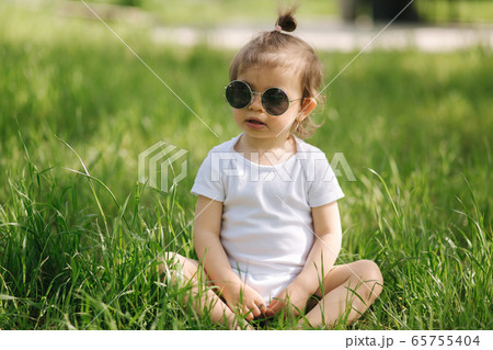 Happy little girl sits on the grass outdoors. Cute baby girl in white bodysuit and sunglasses on the backyard. Adorable kid play with grass 65755404
