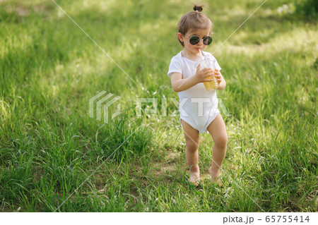 Cute little girl in white bodysuit and sunglasses drink lemonade on the backyard Cute little girl in white bodysuit and sunglasses drink lemonade on the backyard 65755414