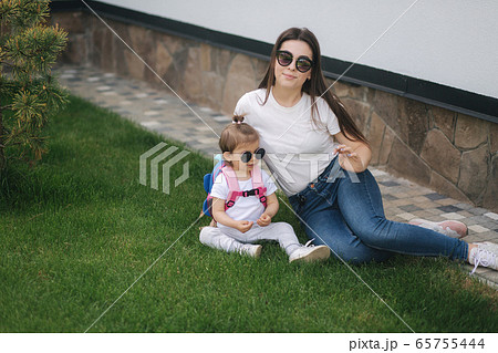 Mom with her adorable little daughter sits on the grass in front of house. Happy family spent time outdoors. Mother and daughter in sunglasses 65755444