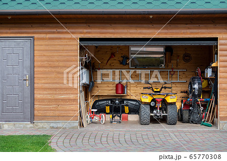 Facade front view open door ATV quad bike motorcycle parking messy garage building with wooden siding at home driveway backyard and lawn path. House warehouse for tools and equipment . Garage sale Facade front view open door ATV quad bike motorcycle parking messy garage building with wooden siding at home driveway backyard and lawn path. House warehouse for tools and equipment . Garage sale 65770308
