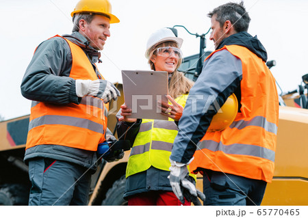 Three workers in a quarry discussing in front of heavy machinery 65770465