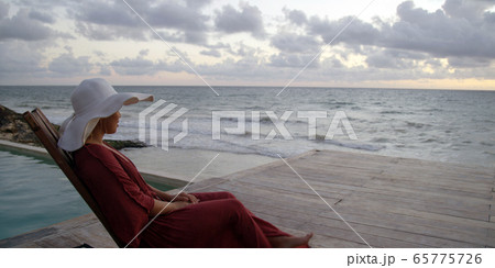 Beautiful young woman sits by the ocean taking in sunset while on vacation Beautiful young woman sits by the ocean taking in sunset while on vacation 65775726