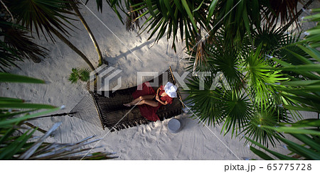 Woman rests in loose red summer dress and hat hanging in hammock surrounded by palm trees at sunset Woman rests in loose red summer dress and hat hanging in hammock surrounded by palm trees at sunset 65775728