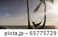 Woman lies in hammock wearing summer dress and hat watching sunset with ocean in the distance 65775729