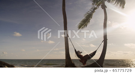 Woman lies in hammock wearing summer dress and hat watching sunset with ocean in the distance 65775729