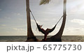 Woman lies in hammock wearing summer dress and hat watching sunset with ocean in the distance 65775730