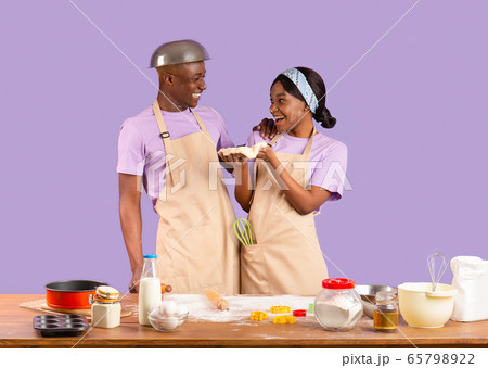 Cheerful black girlfriend and boyfriend enjoying their baking activity on lilac background Cheerful black girlfriend and boyfriend enjoying their baking activity on lilac background 65798922