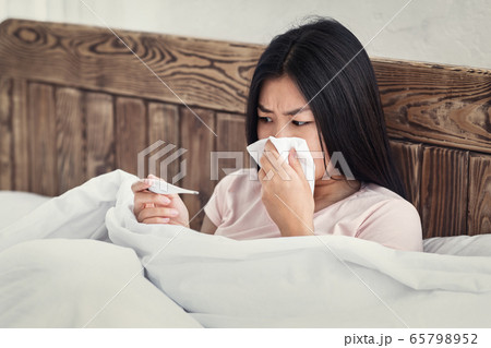 Woman Holding Thermometer Blowing Nose In Tissue Sitting In Bed 65798952