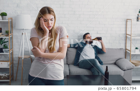 Unhappy woman presses her hand to cheek, man watches TV and drinks beer from bottle at home Unhappy woman presses her hand to cheek, man watches TV and drinks beer from bottle at home 65801794