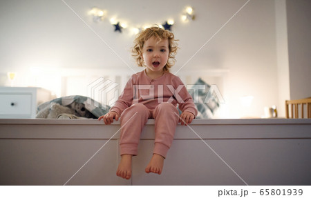 Small toddler girl sitting on bed indoors at home, looking at camera. Small toddler girl sitting on bed indoors at home, looking at camera. 65801939