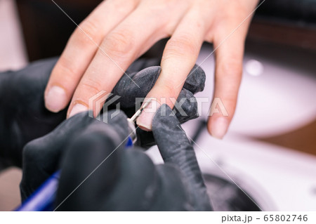 Close-up professional beautician hands working with electric drill on client fingernails. Procedure applying artificial fingernails. Hands of manicurist with tool. Manicurist in gloves removes cuticle Close-up professional beautician hands working with electric drill on client fingernails. Procedure applying artificial fingernails. Hands of manicurist with tool. Manicurist in gloves removes cuticle 65802746