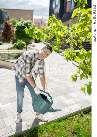 Smiling dark-haired male in protective gloves watering grass with green watering can in the yard Smiling dark-haired male in protective gloves watering grass with green watering can in the yard 65809362