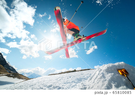 A young athlete skier does a trick on a snow kicker jump and cross skis in a counter light against a blue sky and clouds in the mountains. 65810085