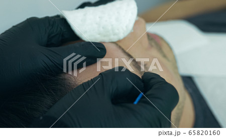 Close up of cosmetologist making permanent makeup. Microblading procedure. High quality close-up of a man laying on a couch during the esthetician brow microblading treatment. Close up of cosmetologist making permanent makeup. Microblading procedure. High quality close-up of a man laying on a couch during the esthetician brow microblading treatment. 65820160