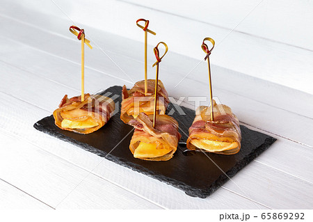 Ravioli wrapped in fried bacon slices, canape on black stone plate, white wooden table. 65869292