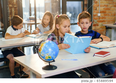 Handsome boy and pretty girl of school sitting together at the desk, look each other and smile while reading book on lesson. Education and pupils concept. 65889594