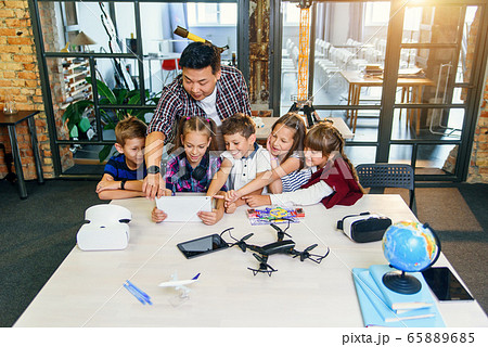 School teacher at desk works with five young pupils using digital tablet computer in technology class. Development, technology and modern education concept. School teacher at desk works with five young pupils using digital tablet computer in technology class. Development, technology and modern education concept. 65889685