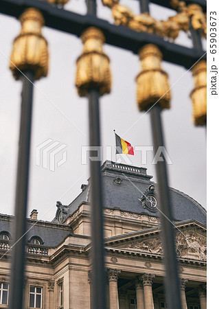 The Royal Palace in Brussels, Belgium. View through the metal fence with golden details. National flag of the Kingdom of Belgium waving on top. The Royal Palace in Brussels, Belgium. View through the metal fence with golden details. National flag of the Kingdom of Belgium waving on top. 65903673