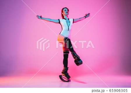 Beautiful redhead woman in a white sportswear jumping in a kangoo jumps shoes isolated on gradient studio background in neon light Beautiful redhead woman in a white sportswear jumping in a kangoo jumps shoes isolated on gradient studio background in neon light 65906305