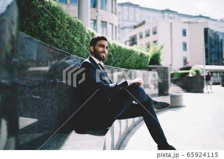 Businessman sitting on bench next to business center and smiling away Businessman sitting on bench next to business center and smiling away 65924115