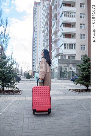 Brunette female in beige coat walking with red suitcase along the street Brunette female in beige coat walking with red suitcase along the street 65941673