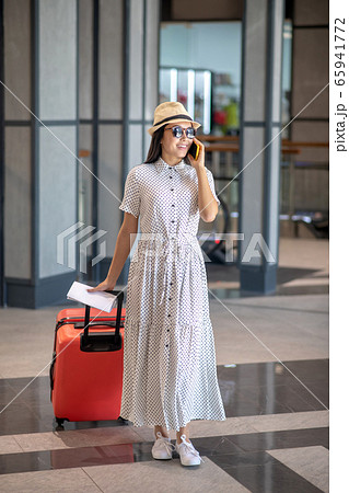 Brunette female in straw hat and sunglasses walking with red suitcase, talking on her smartphone, smiling Brunette female in straw hat and sunglasses walking with red suitcase, talking on her smartphone, smiling 65941772