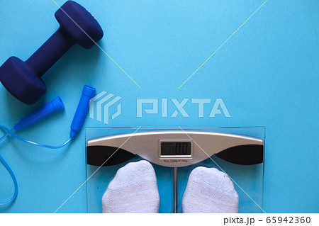 Woman measures her weight by standing her feet in socks on scale on a blue background with skipping rope and dumbbells. Flat lay composition. Healthy active lifestyle Woman measures her weight by standing her feet in socks on scale on a blue background with skipping rope and dumbbells. Flat lay composition. Healthy active lifestyle 65942360