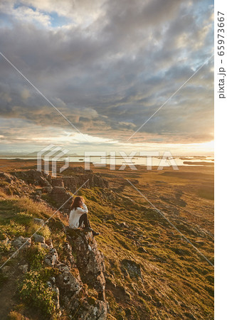 Young woman, female traveler sitting on the top of mountain at sunset. Cloudy dramatic sky. Enjoying wonderful nature in Iceland Young woman, female traveler sitting on the top of mountain at sunset. Cloudy dramatic sky. Enjoying wonderful nature in Iceland 65973667