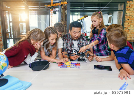In physics and mechanics lesson young Asian teacher demonstrates quadcopter for caucasian pupils at class in modern smart school. Science, drone, engineering and future concept. 65975991