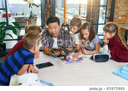 In physics and mechanics lesson young Asian teacher demonstrates quadcopter for caucasian pupils at class in modern smart school. Science, drone, engineering and future concept. In physics and mechanics lesson young Asian teacher demonstrates quadcopter for caucasian pupils at class in modern smart school. Science, drone, engineering and future concept. 65975993