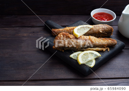 Fried fish hake Pollock with tomato sauce and slices of fresh lemon. Concept eating fatty fast food. Dark wooden background. Copy space. 65984090