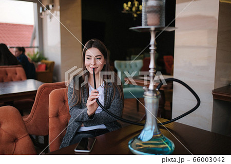 Beautiful young woman in grey jaket smoke hookah on terrace in restautant 66003042