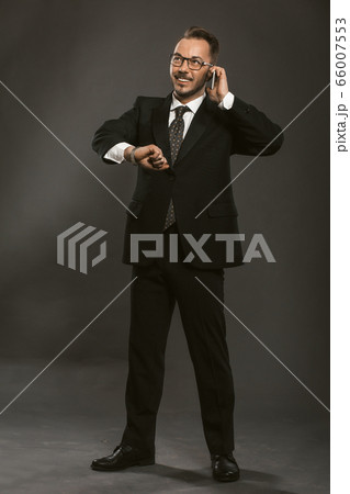 Waiting businessman talks on mobile phone checking time while looking on his wristwatch. Confident well-dressed man smile standing full length on grey background. Toned image Waiting businessman talks on mobile phone checking time while looking on his wristwatch. Confident well-dressed man smile standing full length on grey background. Toned image 66007553