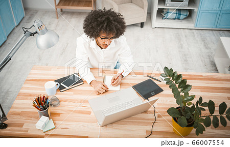 Young copywriter or designer makes draft of new project sitting at wooden table with mobile phone and computer on it. Creative man at home workplace. Planning concept. High angle view. Toned image 66007554
