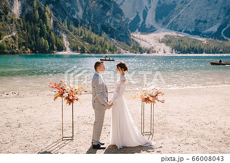 The groom puts on a ring to the bride, at the venue for ceremony, with an arch of autumn flower columns, against the backdrop of Lago di Braies in Italy. Destination wedding in Europe, on Braies lake The groom puts on a ring to the bride, at the venue for ceremony, with an arch of autumn flower columns, against the backdrop of Lago di Braies in Italy. Destination wedding in Europe, on Braies lake 66008043