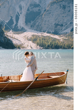 Bride and groom sailing in wooden boat, with oars at Lago di Braies lake in Italy. Wedding in Europe - Newlyweds are standing embracing in a boat. 66008181