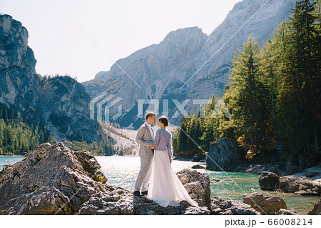 The bride and groom are standing on stones overlooking the Lago di Braies in Italy. Destination wedding in Europe, on Braies lake. Loving newlyweds walk against the backdrop of amazing nature. 66008214