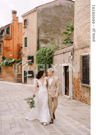Italy wedding in Venice. The bride and groom walk along the deserted streets of the city. The newlyweds hug, dance, hold hands against the backdrop of picturesque red brick houses. Italy wedding in Venice. The bride and groom walk along the deserted streets of the city. The newlyweds hug, dance, hold hands against the backdrop of picturesque red brick houses. 66008391