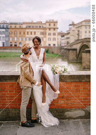 Interracial wedding couple. Wedding in Florence, Italy. An African-American bride is sitting on a brick wall and Caucasian groom is hugging her. Arno River Embankment, overlooking city and bridges Interracial wedding couple. Wedding in Florence, Italy. An African-American bride is sitting on a brick wall and Caucasian groom is hugging her. Arno River Embankment, overlooking city and bridges 66008488
