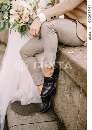 Wedding in Florence, Italy. Close-up of the legs of the groom in light short pants and black leather shoes, the bride in a white dress with a bouquet in her hands. 66008544