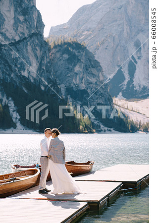 The bride and groom walk along a wooden boat dock at the Lago di Braies in Italy. Wedding in Europe, on Braies lake. Newlyweds walk, kiss, hug on a background of rocky mountains. 66008545