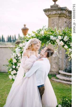 Wedding at an old winery villa in Tuscany, Italy. The groom circles the bride in his arms. Round wedding arch decorated with white flowers and greenery in front of an ancient Italian architecture. Wedding at an old winery villa in Tuscany, Italy. The groom circles the bride in his arms. Round wedding arch decorated with white flowers and greenery in front of an ancient Italian architecture. 66008791