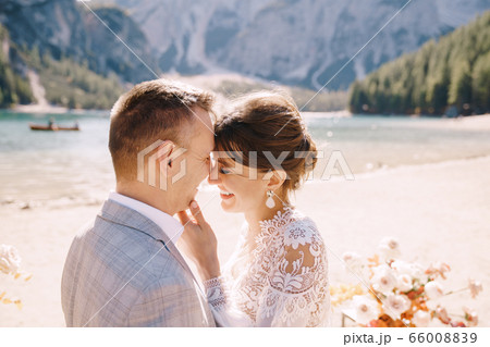 The newlyweds kiss on the spot for the ceremony, with an arch of autumn flower columns, against the backdrop of the Lago di Braies in Italy. Destination wedding in Europe, on Braies lake. 66008839