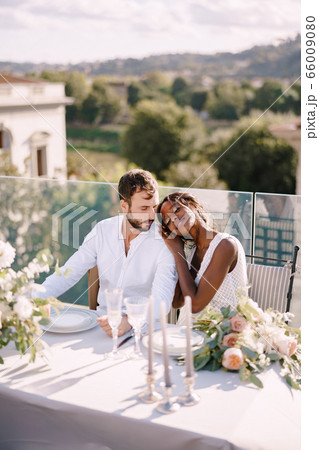 Destination fine-art wedding in Florence, Italy. African-American bride and Caucasian groom are sitting at the rooftop wedding dinner table overlooking the city. Multiracial wedding couple Destination fine-art wedding in Florence, Italy. African-American bride and Caucasian groom are sitting at the rooftop wedding dinner table overlooking the city. Multiracial wedding couple 66009080